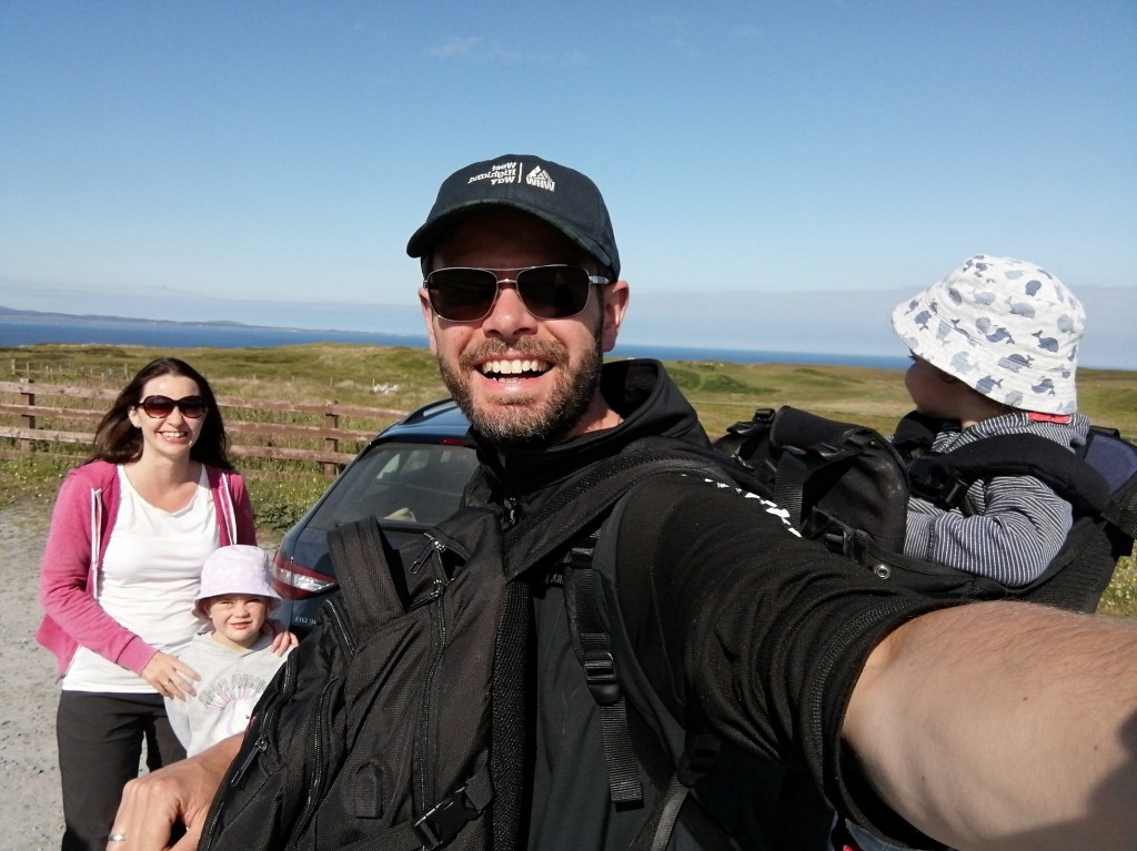guy smiling, baby carrier, sunglasses, blue skies, islay, scotland